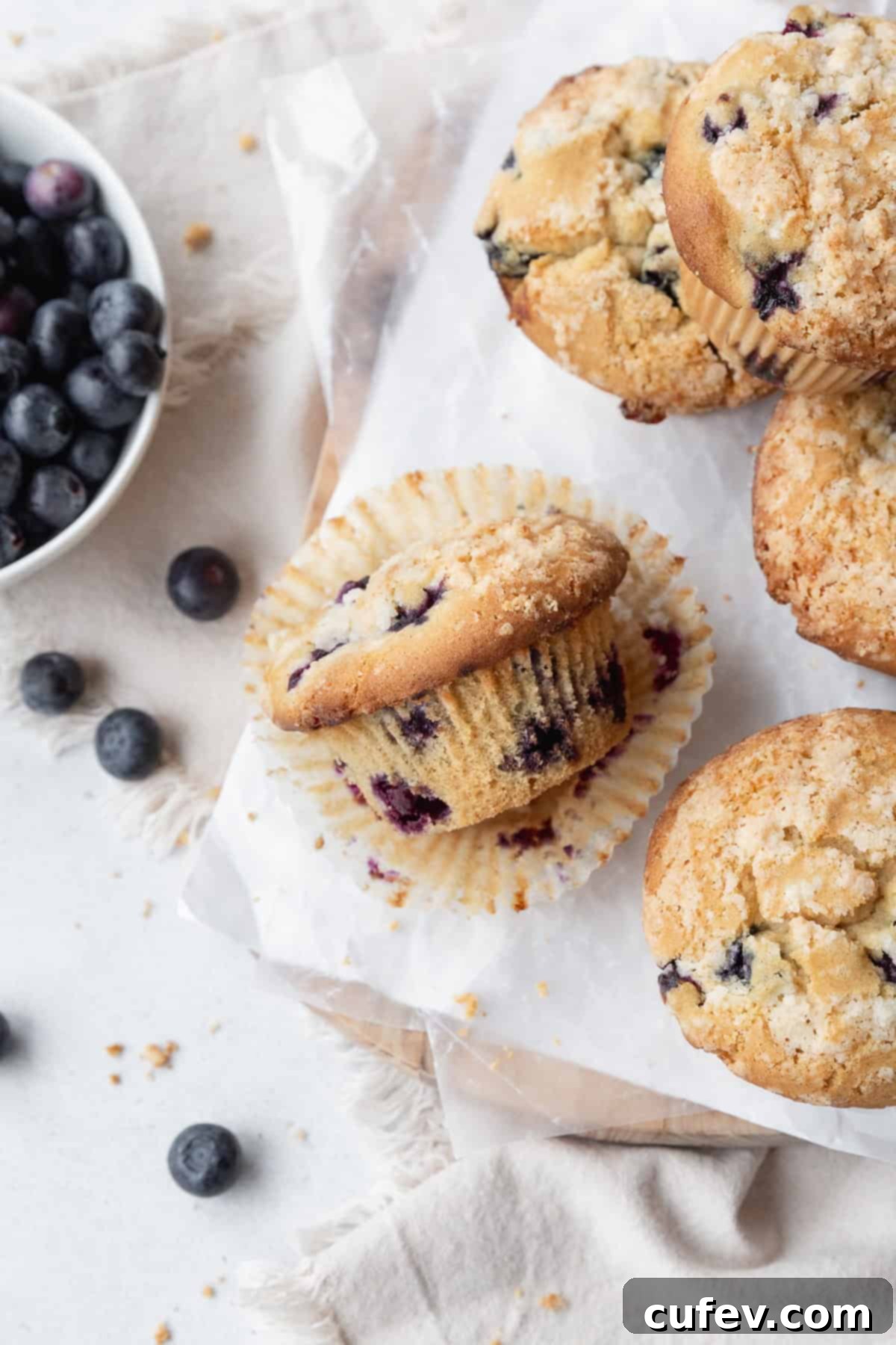 A blueberry muffin laying on its side on a paper liner, with more muffins on the right and a bowl of blueberries on the left.