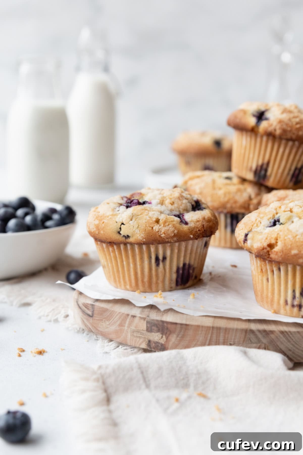Gluten-free blueberry muffins on a wooden board lined with parchment paper, with a bowl of blueberries on the side and milk jars in the background.