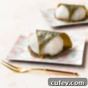 Sakura mochi wrapped in a cherry blossom leaf on a square pink plate with a gold fork laying in front.