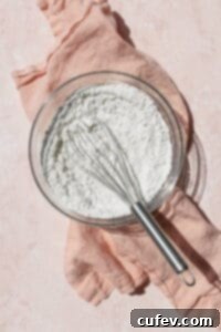 A clear glass bowl filled with a mixture of dry ingredients for waffle batter, including flour, sugar, and baking powder, expertly whisked together on a soft pink surface, ready for the next step of preparation.