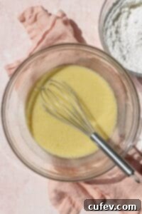 A glass bowl containing the liquid components of waffle batter – eggs, buttermilk, oil, and vanilla – being vigorously whisked together to create a smooth, unified mixture.