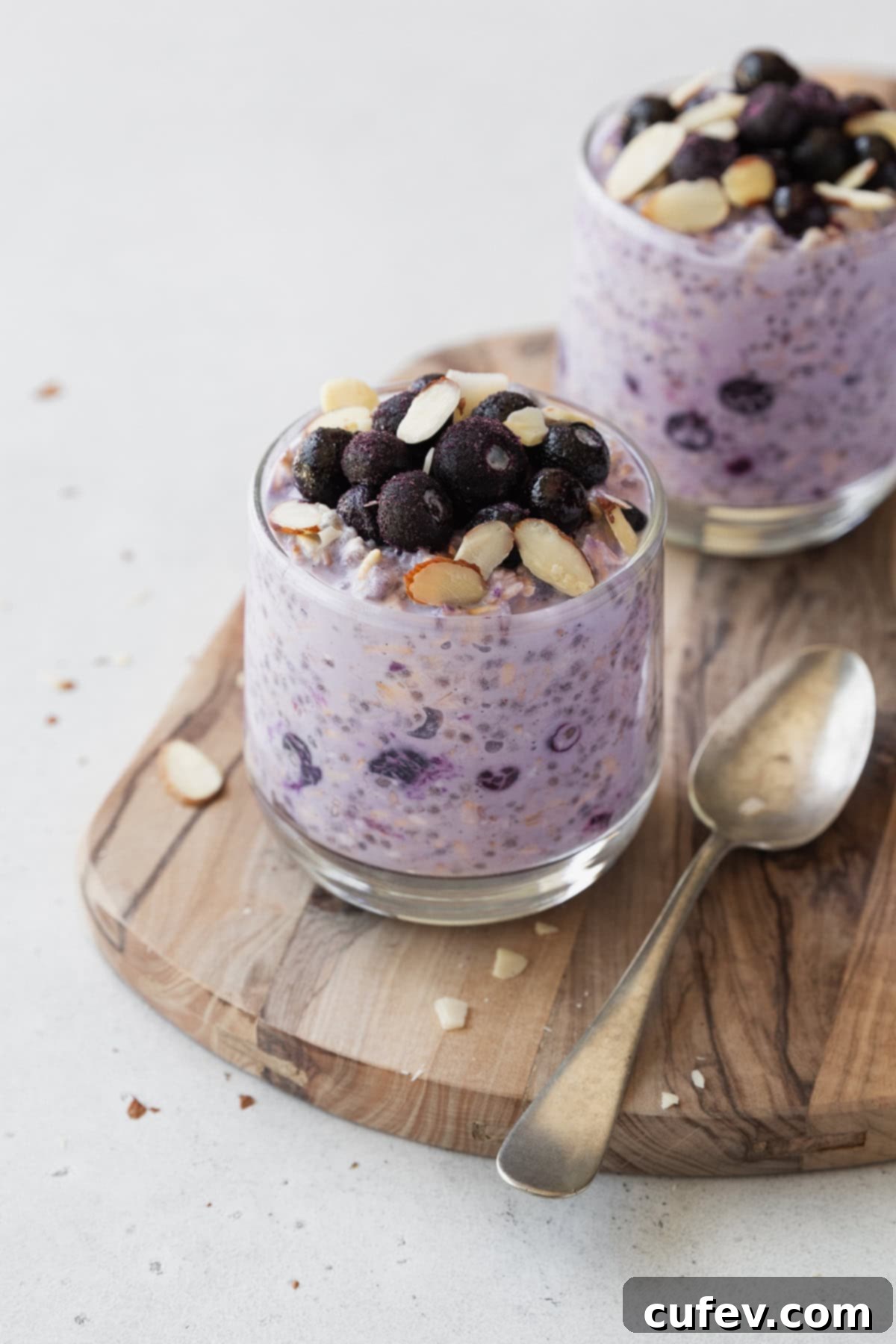 Wholesome Blueberry Overnight Oats 2 Two glass of blueberry overnight oats on a wooden board with a spoon, ready to be enjoyed.