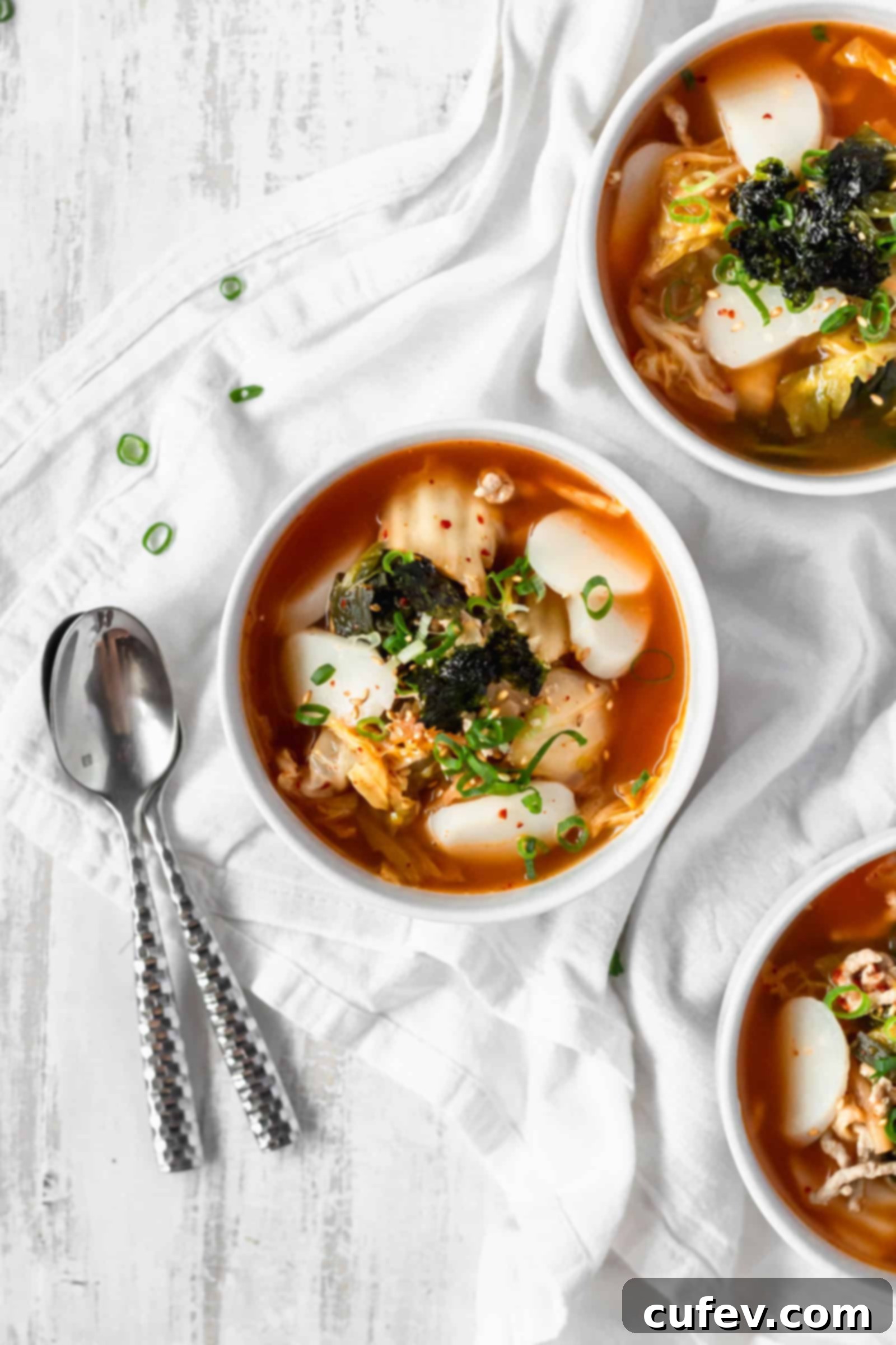 Kimchi Tteokguk A Zesty Korean Comfort Bowl 2 Three bowls of Tteokguk (Korean Rice Cake Soup) on a white surface with two silver spoons, garnished with green onions and seaweed.