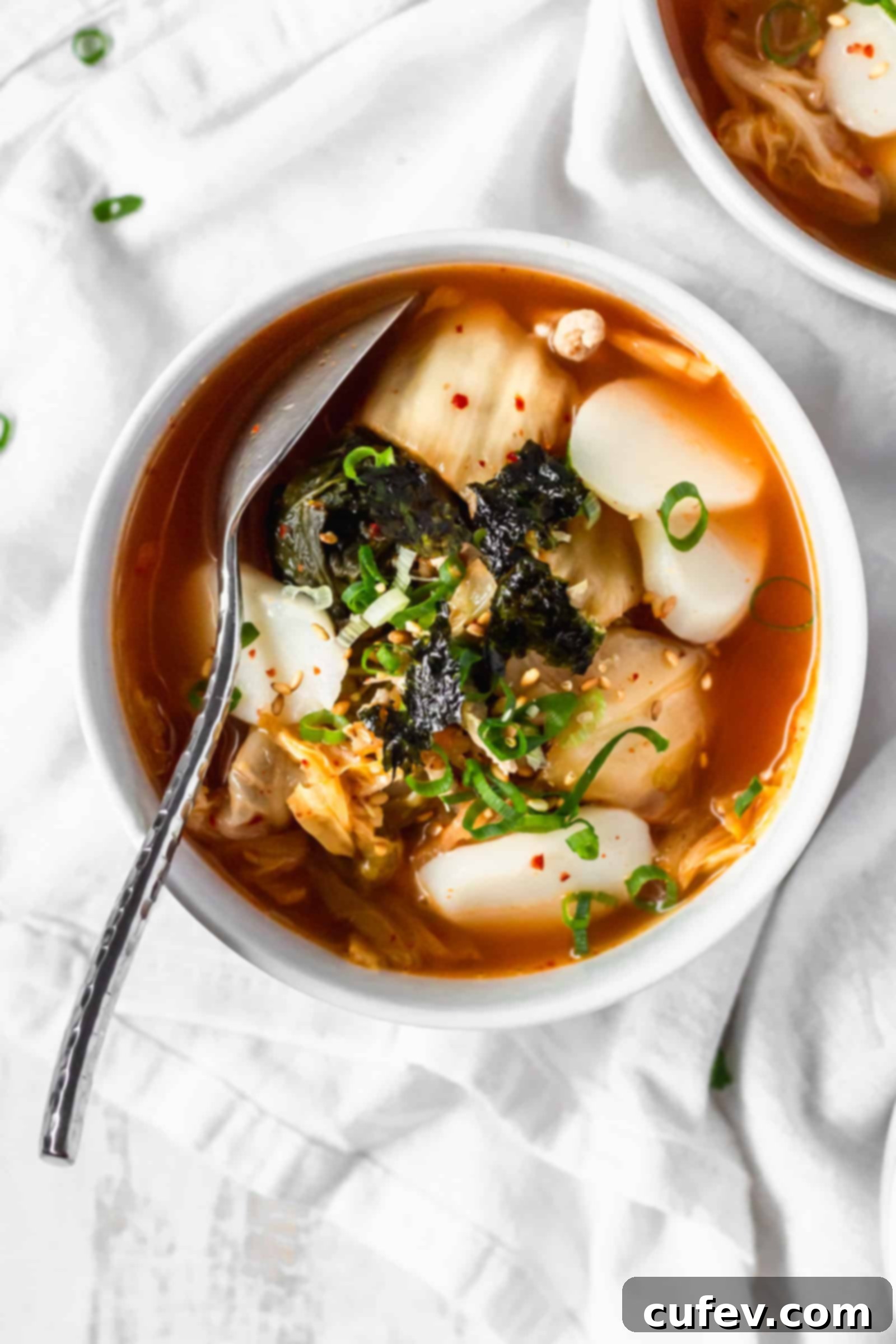 Kimchi Tteokguk A Zesty Korean Comfort Bowl 3 An overhead shot of a steaming bowl of Korean rice cake soup, garnished with kimchi, green onions, and seaweed, on a white surface.