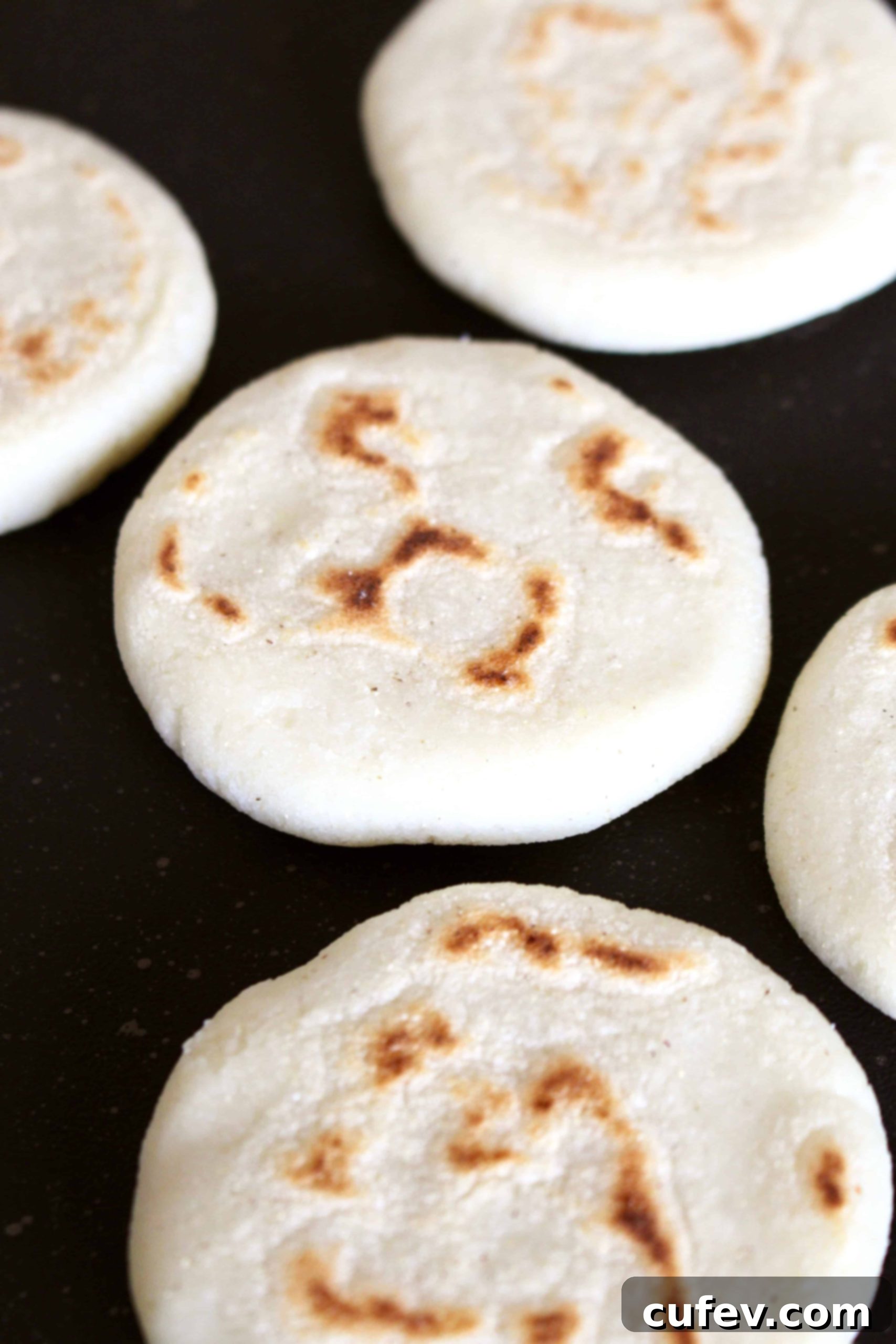 Two freshly shaped arepa patties cooking on a dark metal pan, showing early signs of browning.