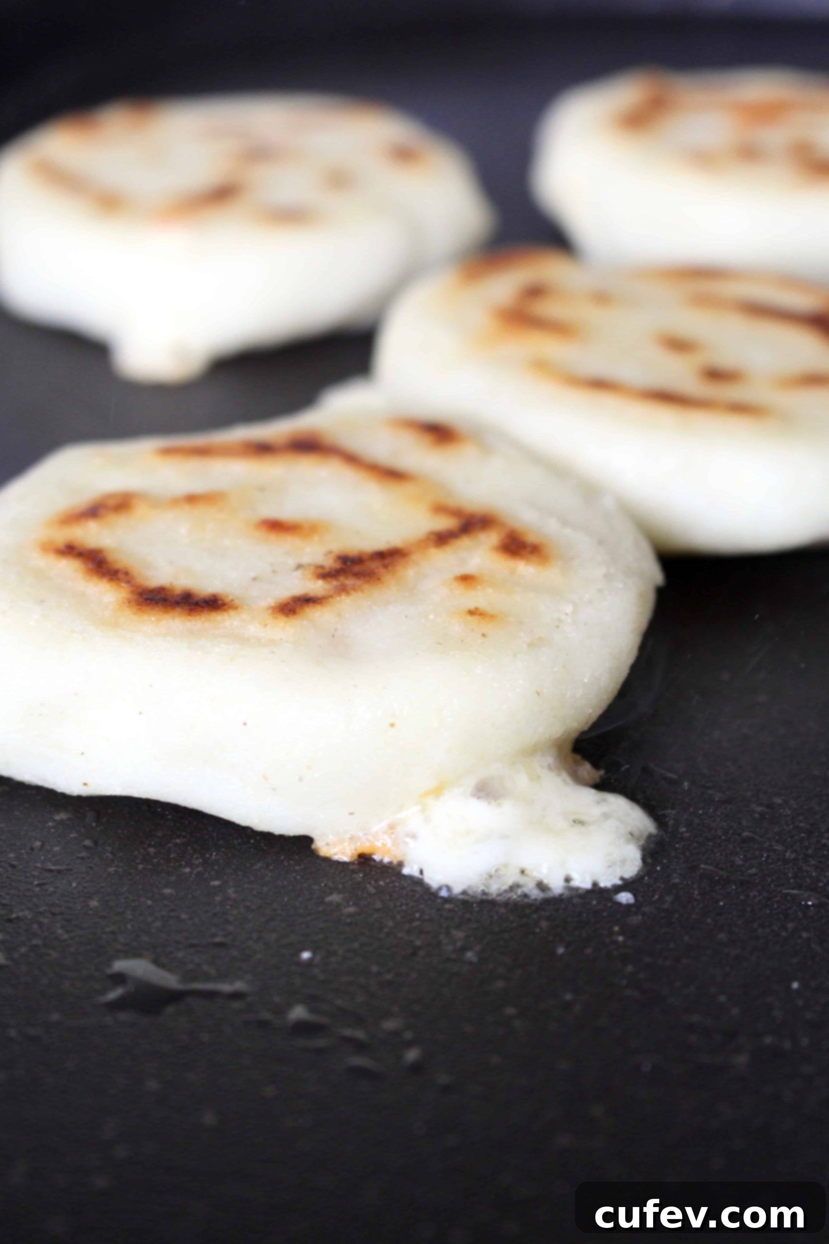A close-up of an arepa cooking on a black griddle, with luscious melted cheese gently oozing out, indicating a perfect cheese-stuffed arepa.