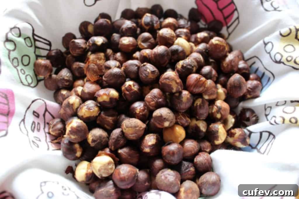 Roasted hazelnuts, still warm, are being poured from a baking sheet into a tea towel-lined bowl.