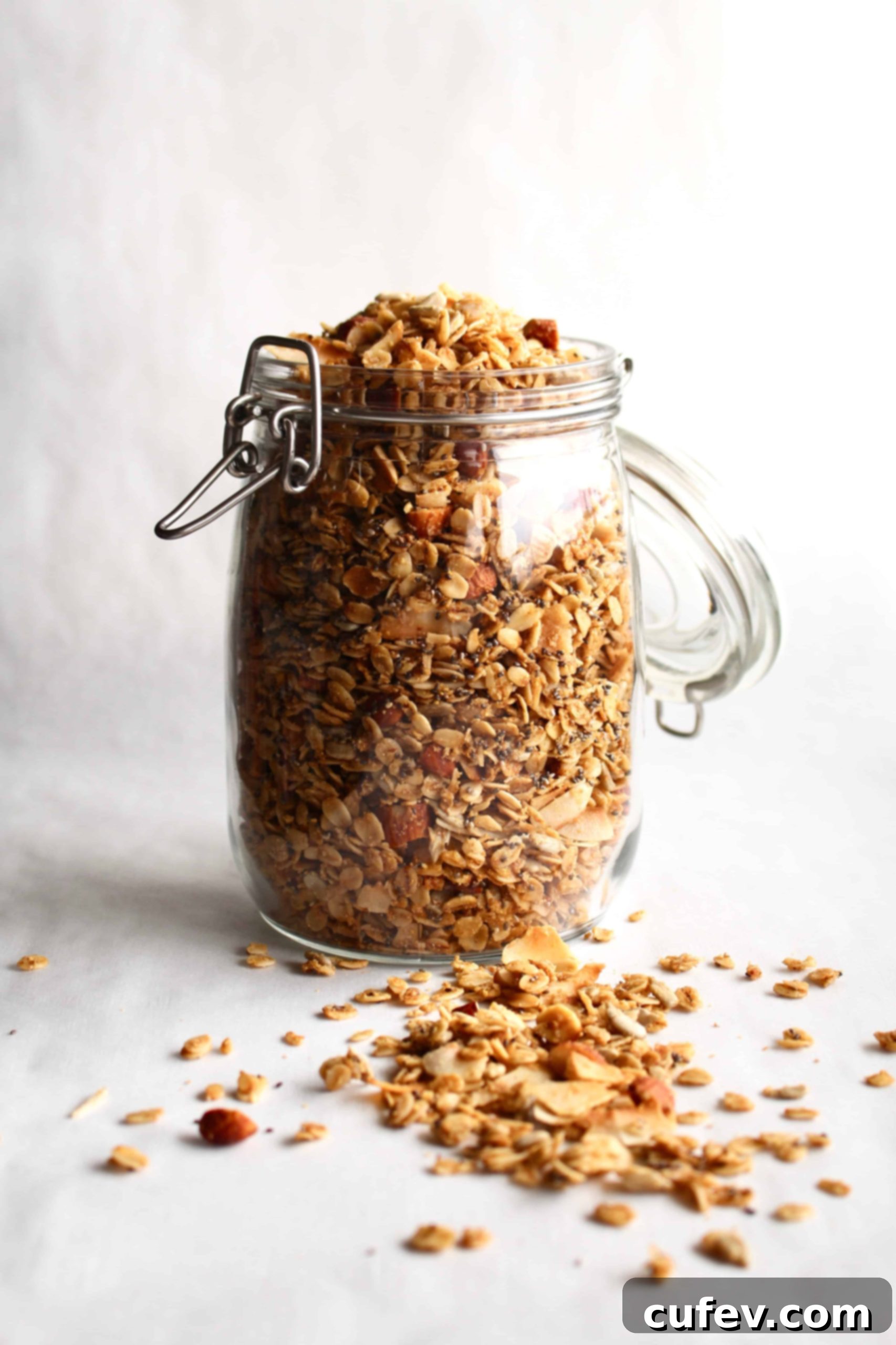 A glass jar of homemade gluten-free granola on a white surface, with some golden-brown granola spilled invitingly in front of the jar.