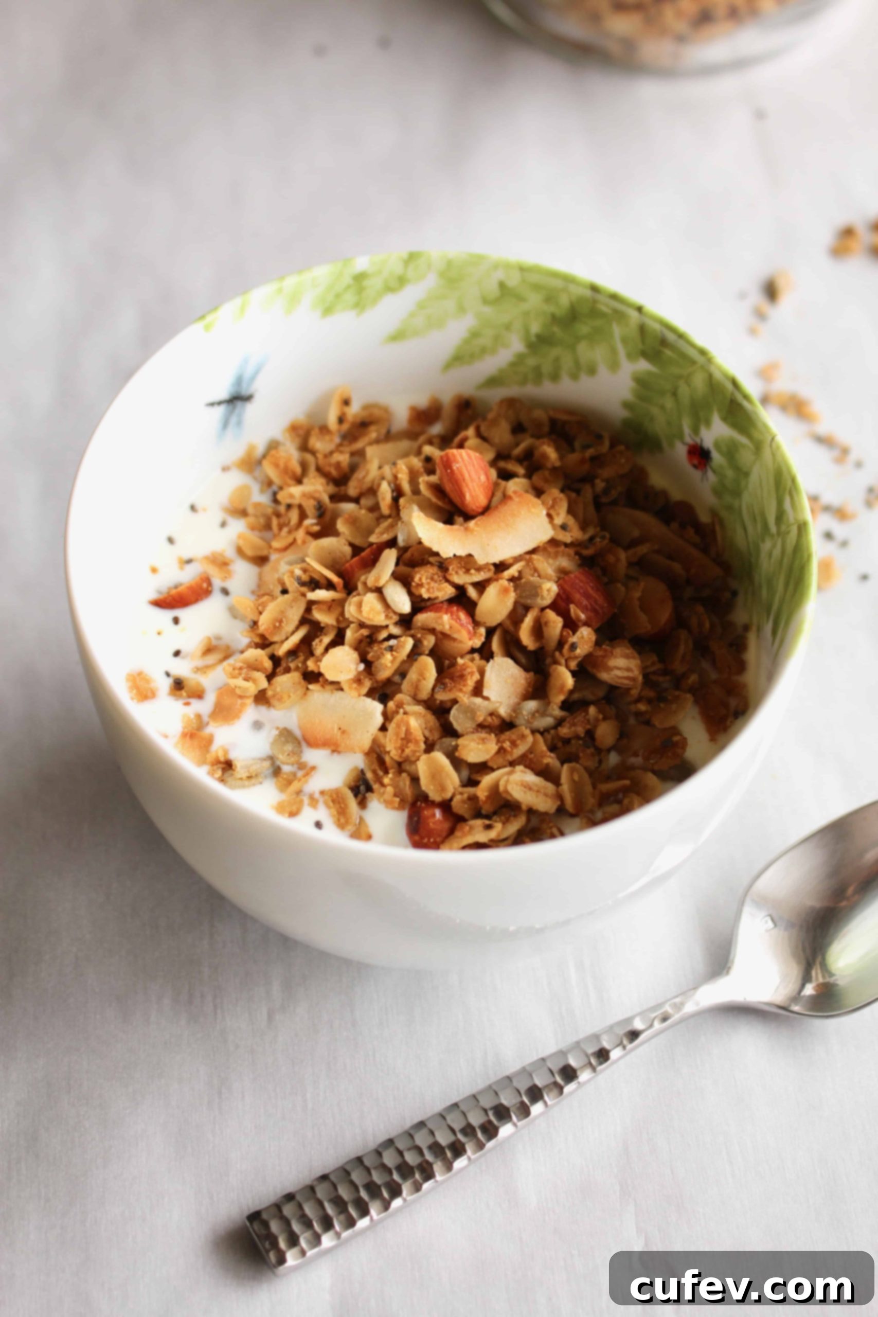 A white bowl filled with crunchy homemade gluten-free granola and a splash of milk, with a silver spoon resting elegantly next to it.