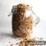 A glass jar of homemade gluten-free granola on a white surface with some spilled out, showcasing its golden crunch.