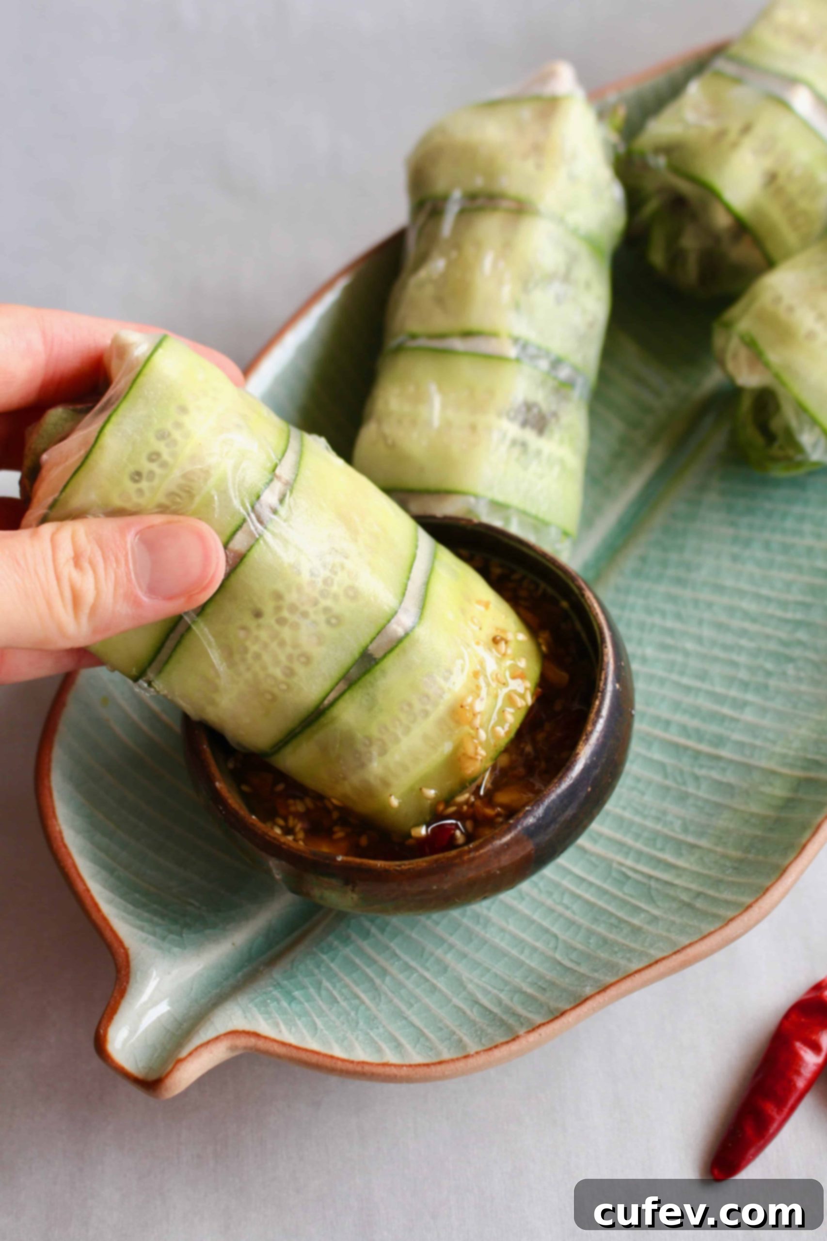 A hand dipping a summer roll with cucumbers into a dish of spicy sauce.