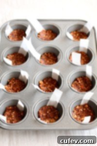 A mini muffin pan showing individual wells filled with a dark, compact vegan crust, ready for the cheesecake filling.