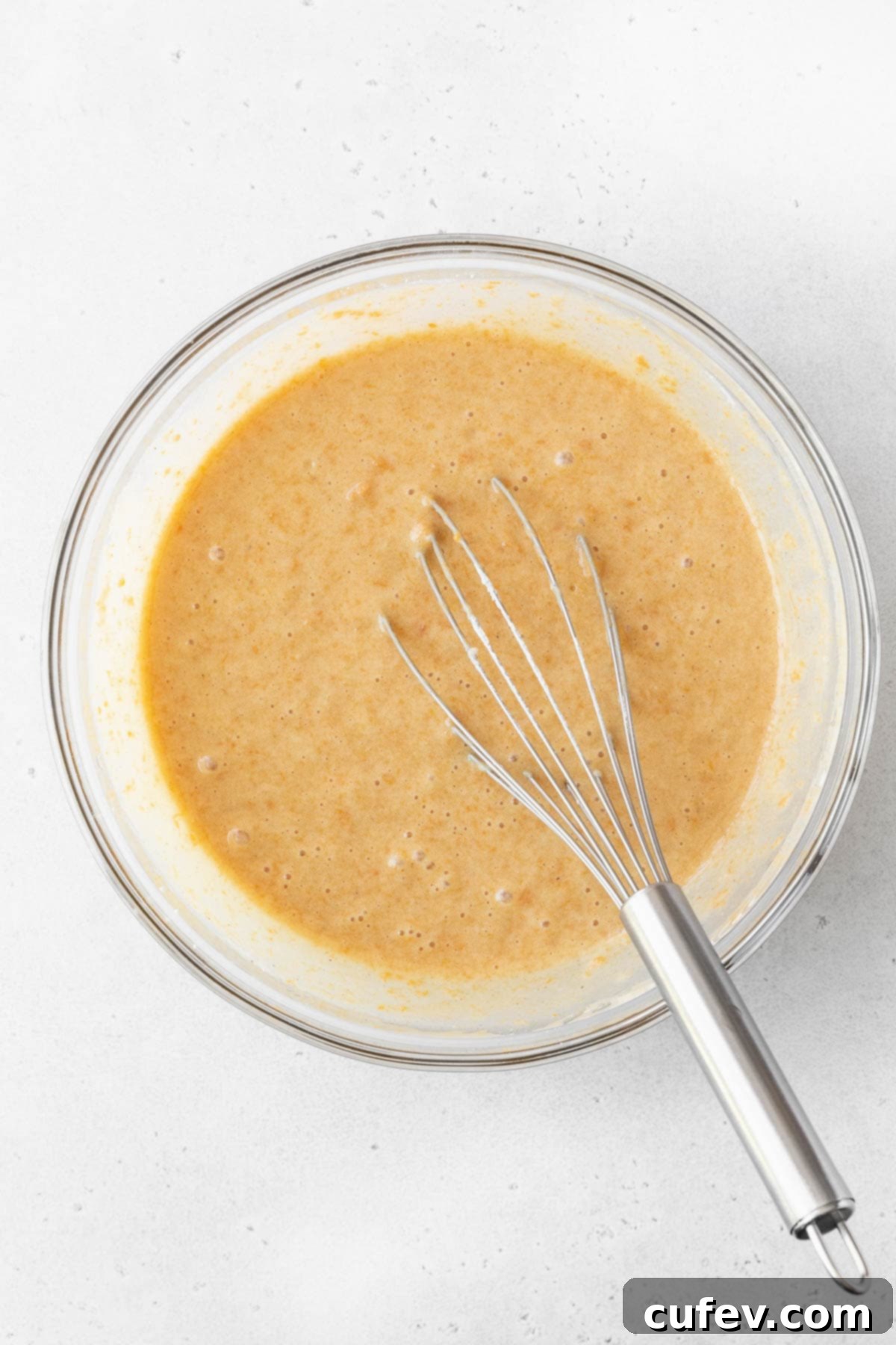 The healthy sweet potato cake batter, perfectly mixed and showcasing its rich color, rests in a glass bowl with a silver whisk nearby.
