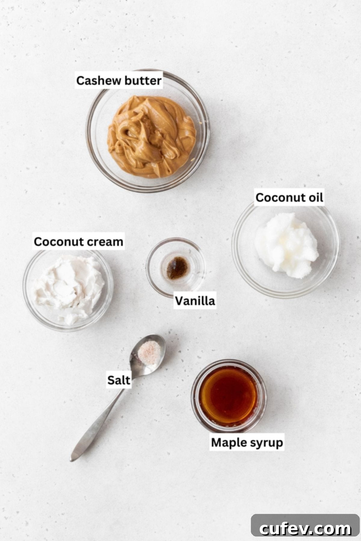 Overhead shot of all the maple cashew frosting ingredients: cashew butter, refined coconut oil, coconut cream, maple syrup, vanilla extract, and fine sea salt, arranged on a white surface.