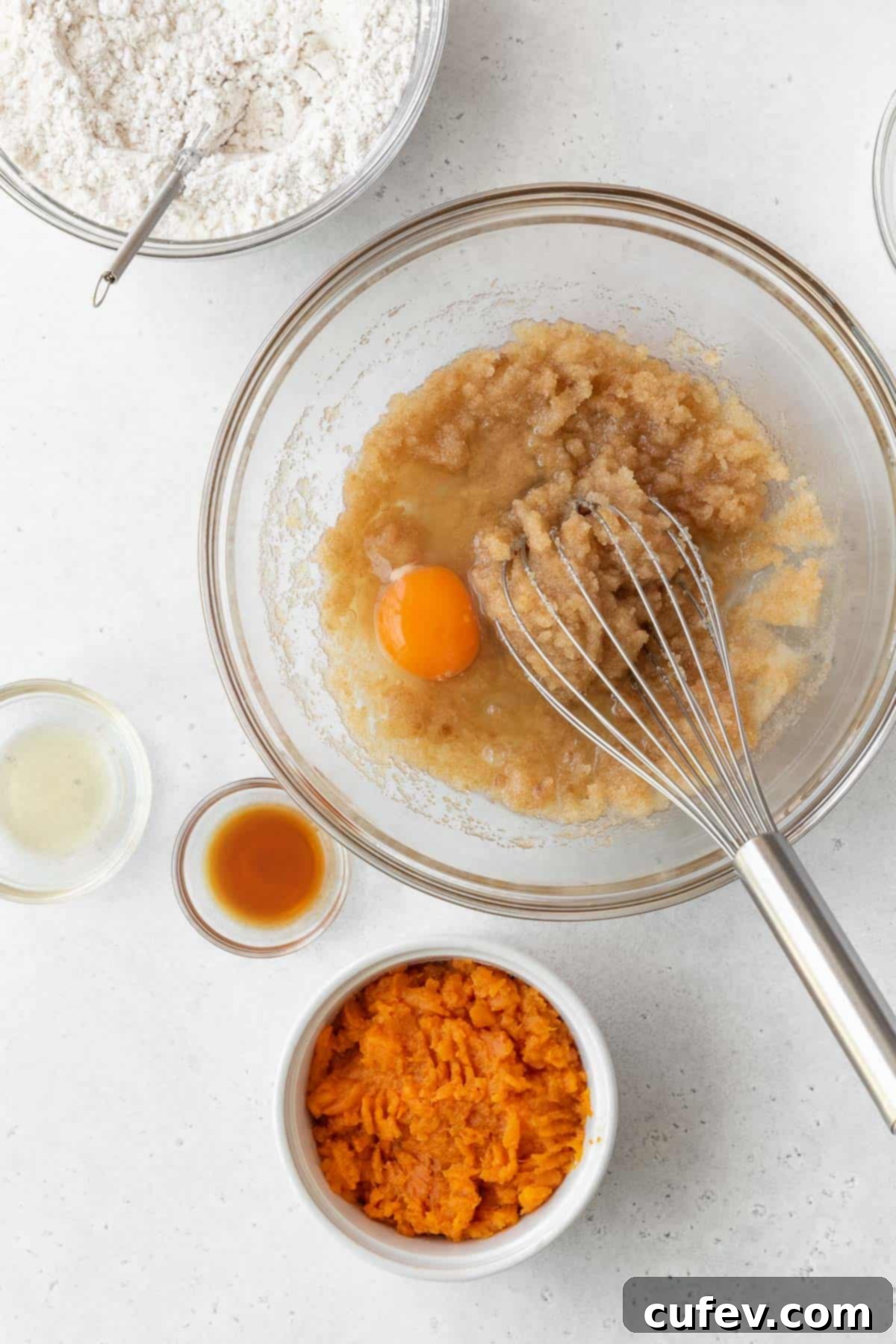 An egg being carefully whisked into the glistening oil and sugar mixture in a glass bowl, ensuring a smooth blend.