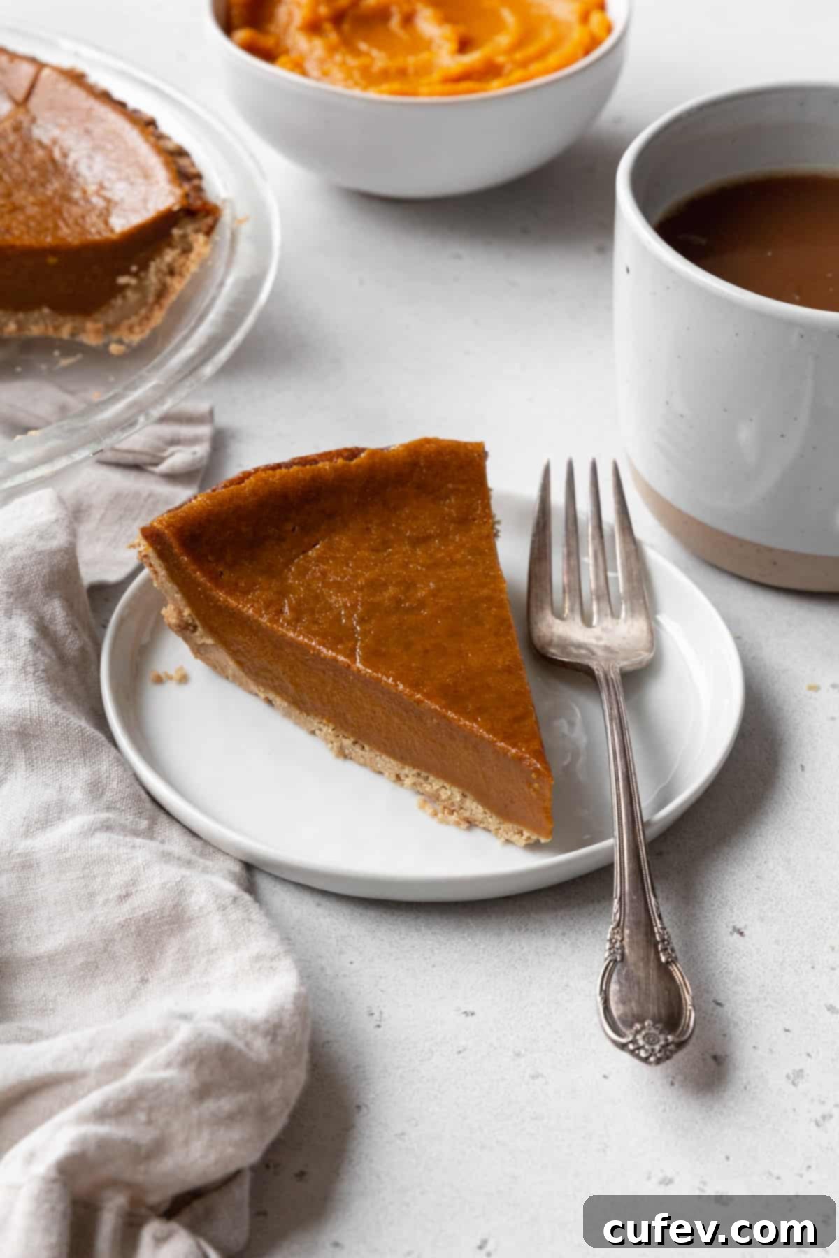 A beautifully presented slice of eggless pumpkin pie on a white dessert plate, accompanied by a silver spoon, with a cozy mug of coffee gently blurred in the background. Perfect for holiday gatherings.