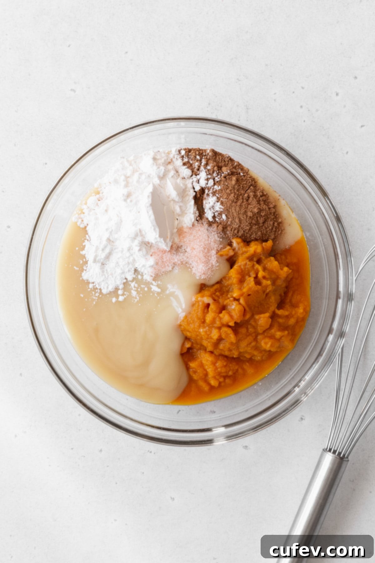 All the vibrant pumpkin pie filling ingredients gathered in a glass bowl, ready to be mixed, with a wire whisk nearby.
