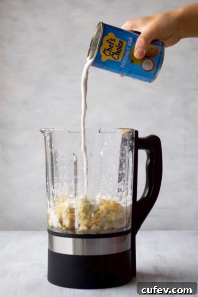 A can of coconut milk being poured into a blender with cooked cauliflower and other ingredients for the creamy sauce.
