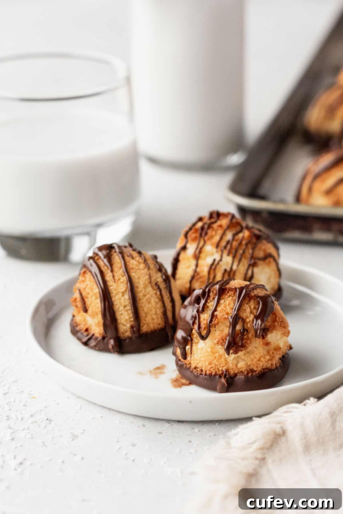 Three chocolate-drizzled coconut macaroons on a white plate, with a glass of plant-based milk in the background, ready to be enjoyed.