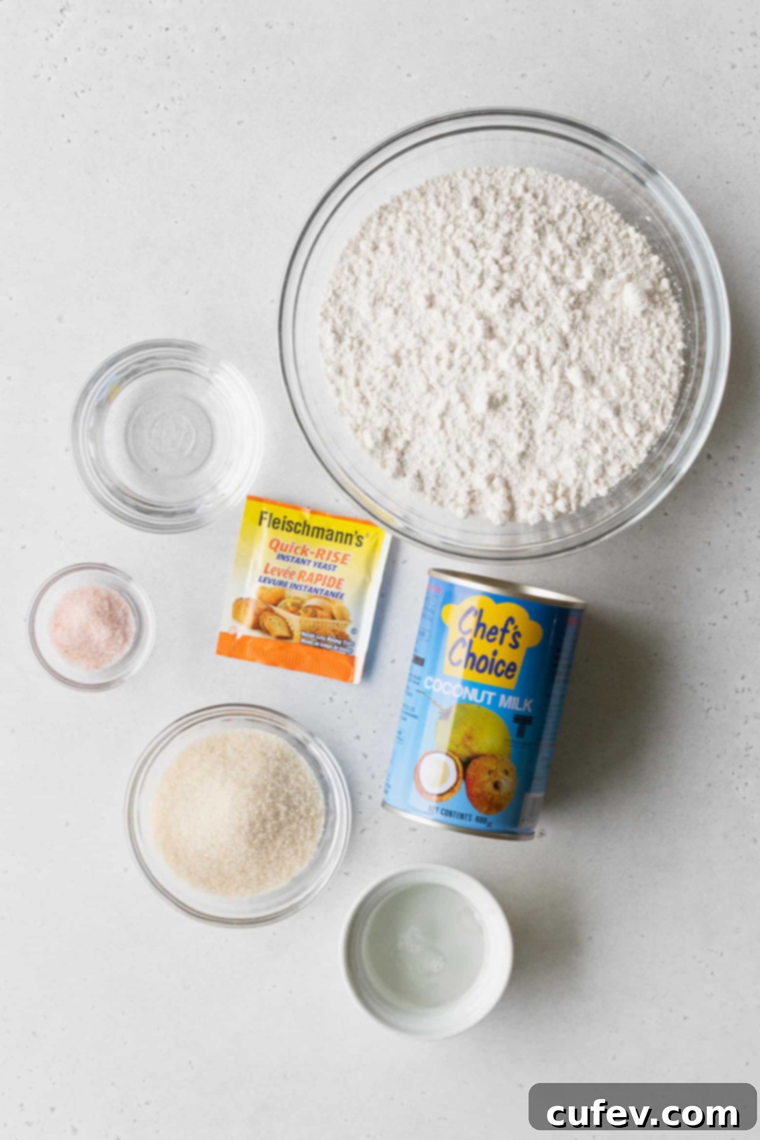 Various ingredients for making pani popo, including flour, sugar, coconut oil, yeast, and a can of coconut milk, precisely measured in bowls.