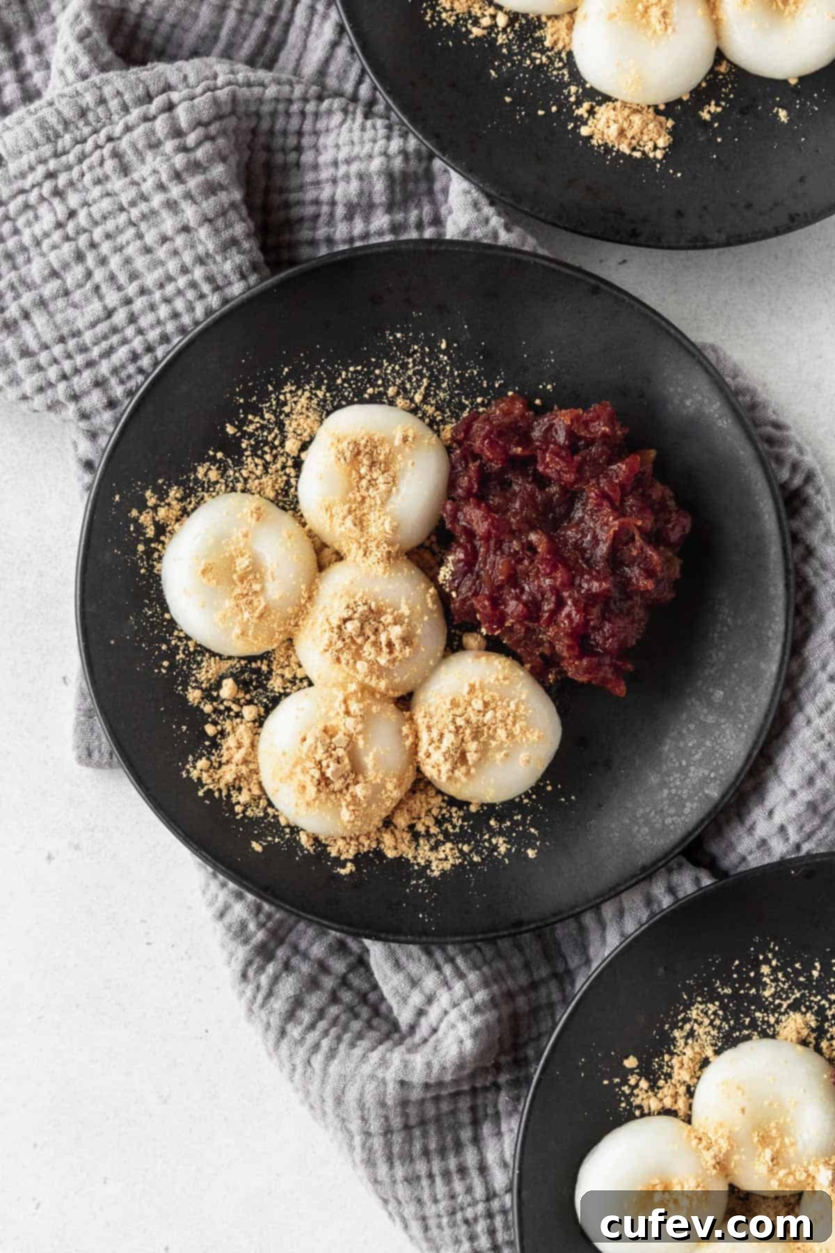 An overhead shot of a black plate with shiratama dango, sweet red bean paste, and kinako sprinkled on top, with two more plates in the corner of the shot.