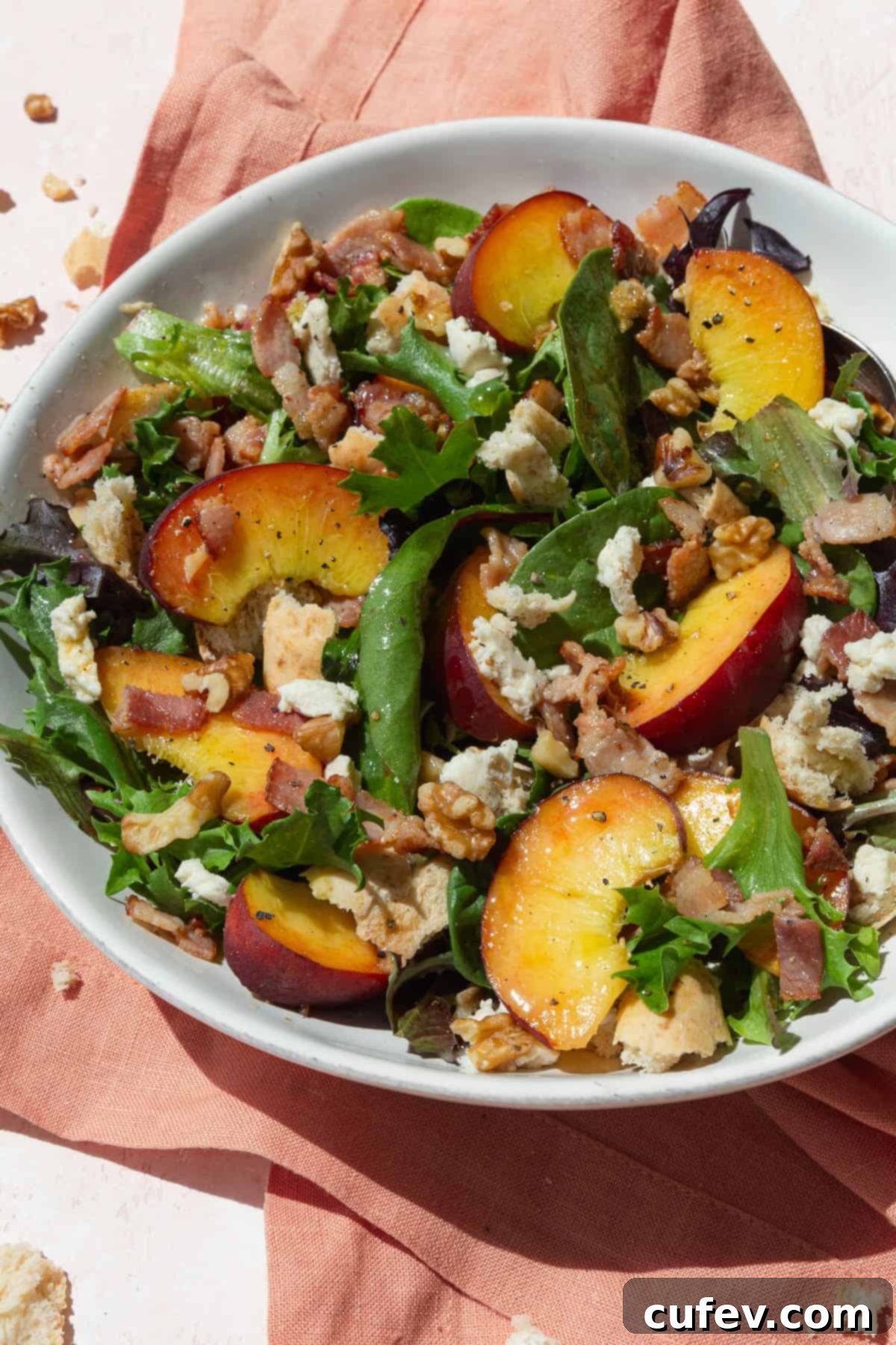 Peach salad with baby greens, cheese, and walnuts in a white bowl on a pink napkin.