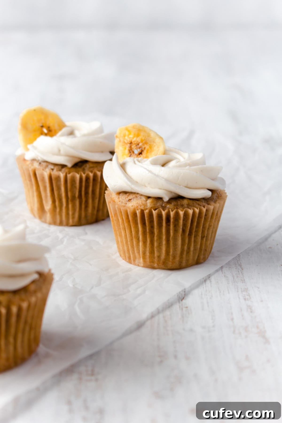 Two banana cupcakes on a piece of parchment paper with another cupcake off to the side, showcasing their perfect dome and inviting texture.