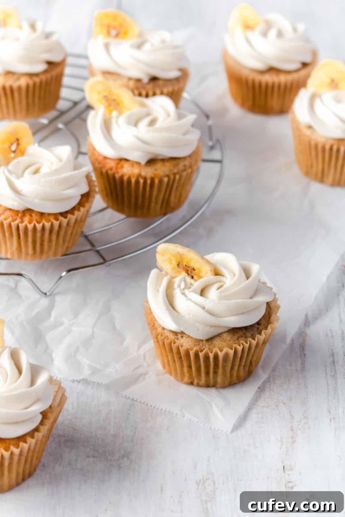 Banana cupcakes with buttercream frosting piped on top and decorated with a banana chip scattered on a white surface, looking perfectly delicious.