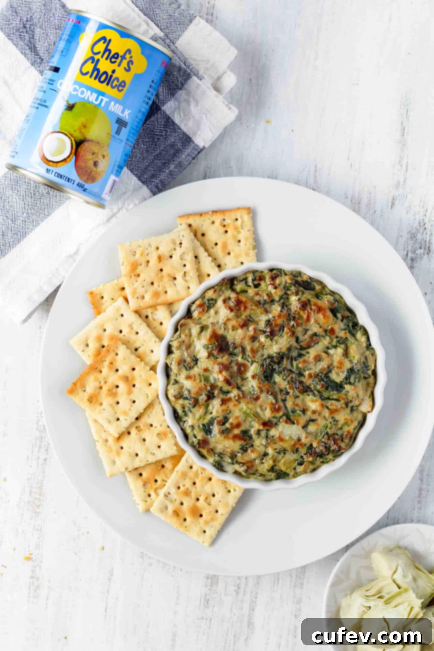 A white plate with a dish of hot vegan spinach artichoke dip and crackers, with a blue can of coconut milk in the soft-focus background.