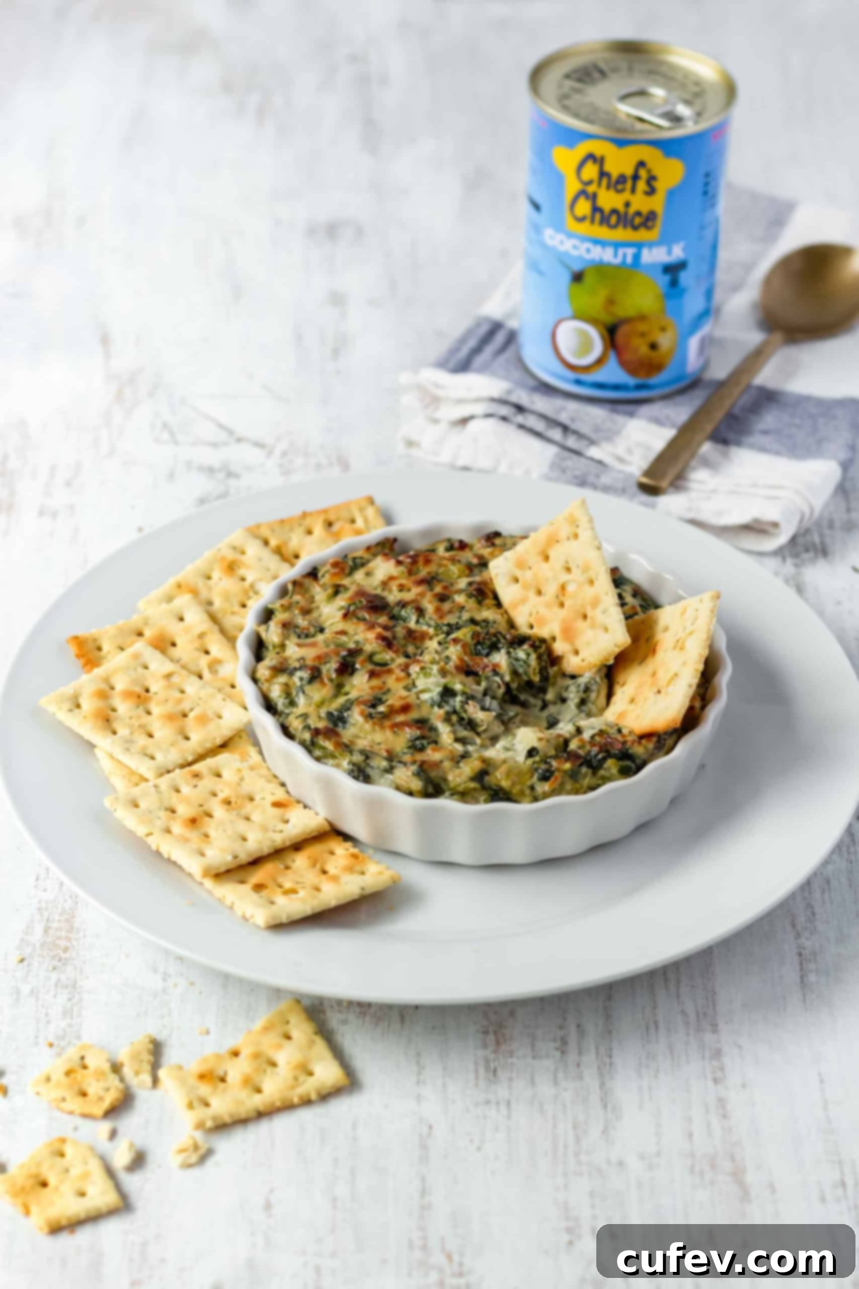A festive arrangement featuring a bowl of golden-brown vegan spinach artichoke dip, accompanied by crunchy crackers and a coconut milk can in the soft-focus background.
