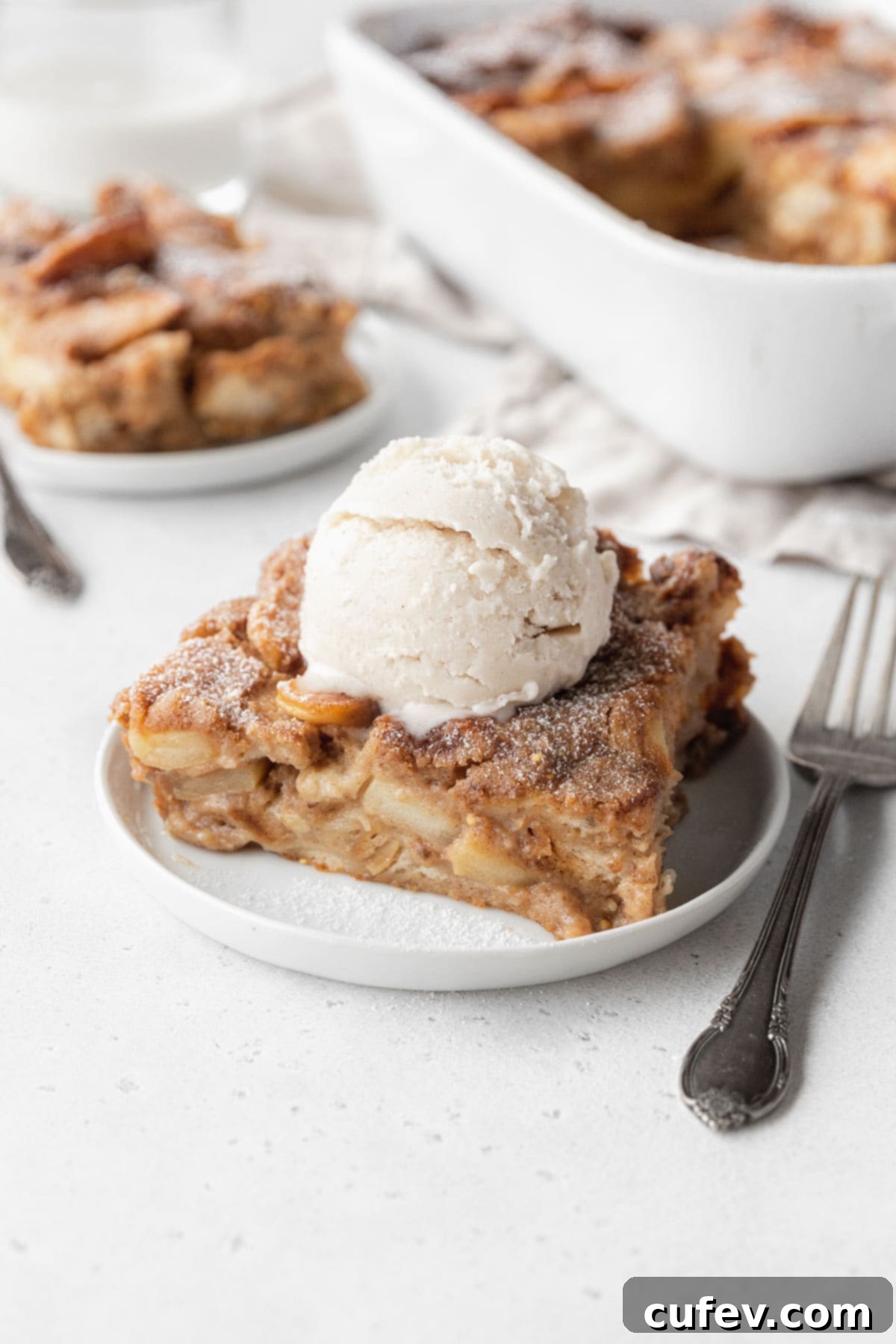 Cozy Vegan Bread Bake 2 A perfectly sliced piece of apple pie vegan bread pudding on a pristine white dessert plate, elegantly topped with a melting scoop of vegan ice cream and a hint of caramel drizzle.