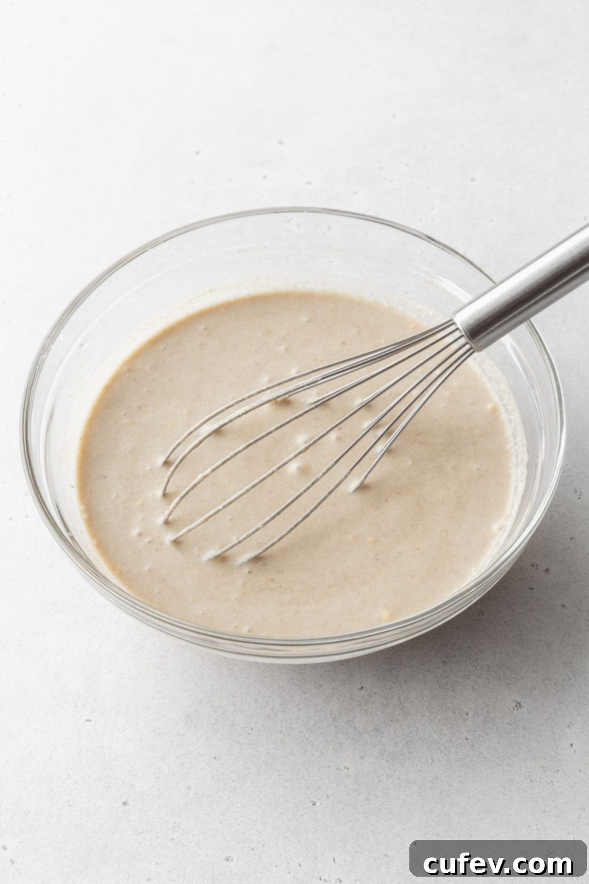 Cozy Vegan Bread Bake 11 A hand whisking smooth vegan bread pudding batter in a clear glass mixing bowl, showing its creamy texture.