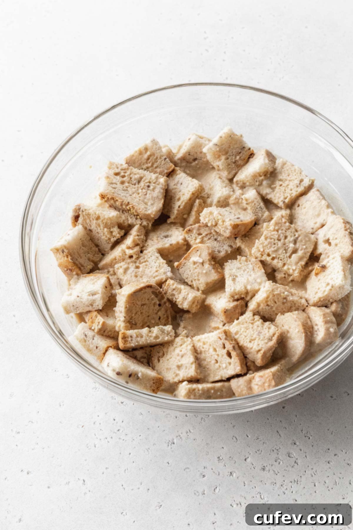 Cozy Vegan Bread Bake 12 Cubed stale bread soaking thoroughly in a creamy, off-white vegan bread pudding batter within a glass bowl, absorbing the liquid.