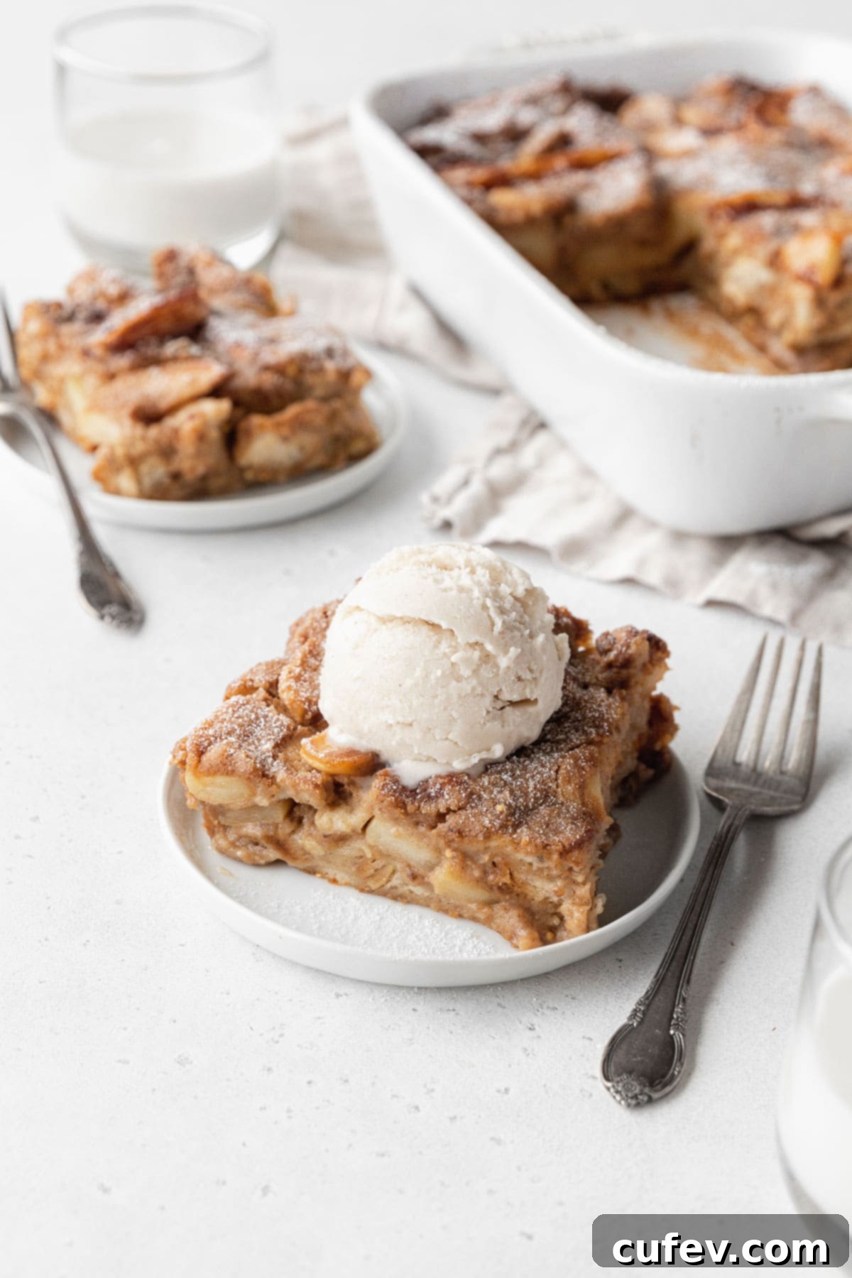 Cozy Vegan Bread Bake 16 A beautifully presented slice of vegan bread pudding on a white plate, topped with a scoop of vegan vanilla ice cream. In the background, another plate and the full white casserole dish are softly blurred, creating a cozy and inviting scene.