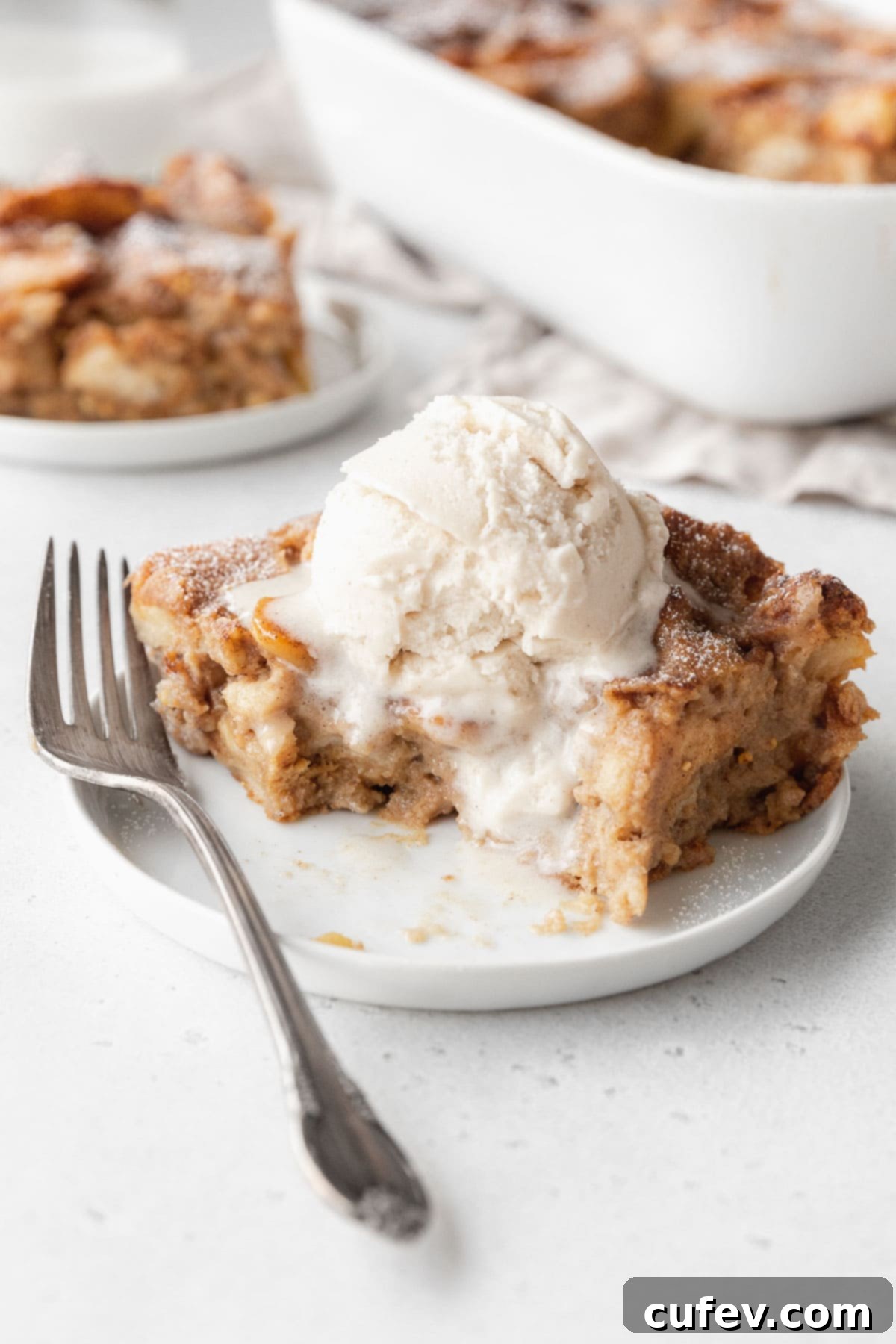 Cozy Vegan Bread Bake 17 A partially eaten slice of vegan bread pudding on a white plate, featuring a melting scoop of vanilla ice cream on top, inviting another delicious bite.