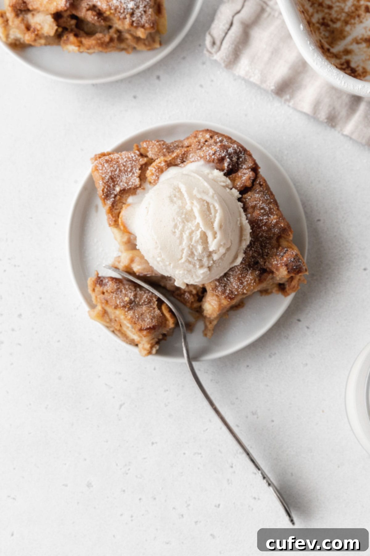Cozy Vegan Bread Bake 4 An inviting overhead shot captures a slice of vegan bread pudding, elegantly topped with a scoop of creamy vanilla ice cream, as a silver fork gently cuts into a corner, hinting at its soft texture.