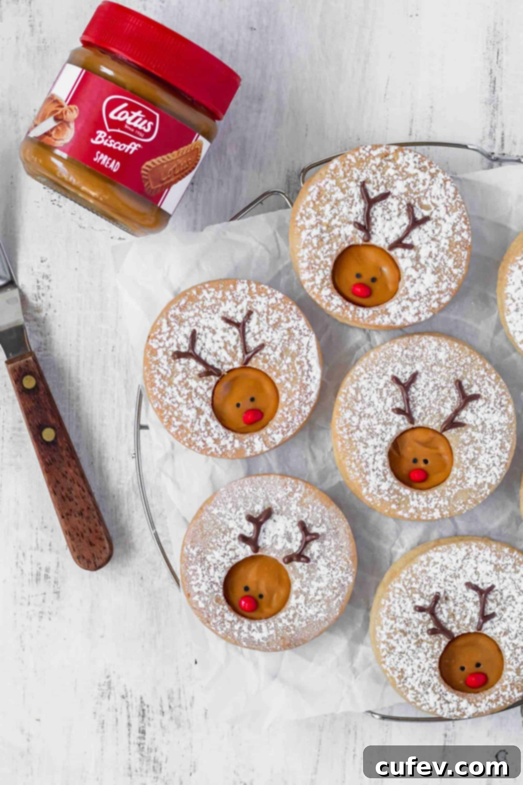 Two Rudolph reindeer cookies resting on parchment paper next to an open jar of Biscoff spread, ready for assembly.
