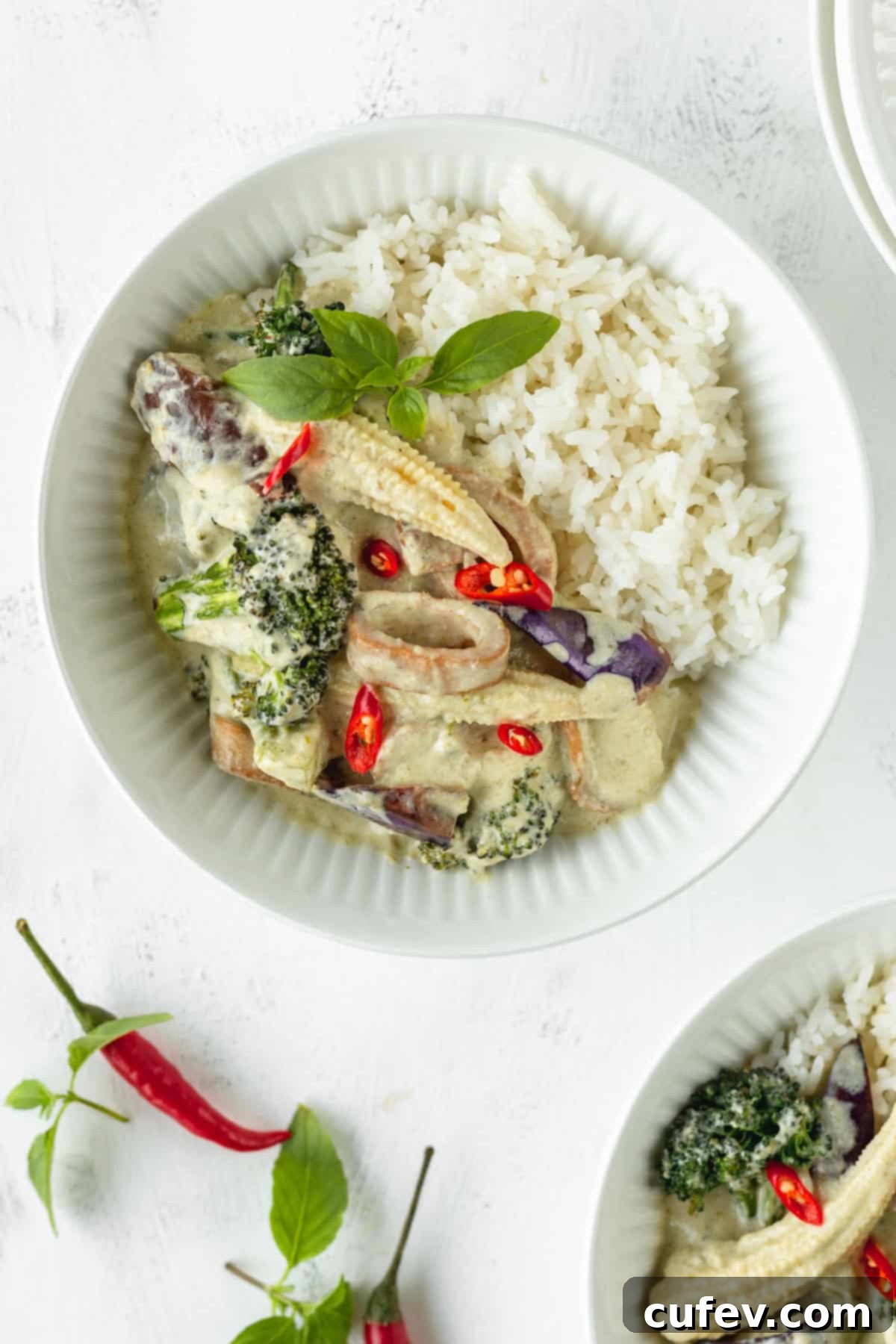 A white bowl of vegan Thai curry with rice on a white surface with some herbs and chili peppers scattered around.