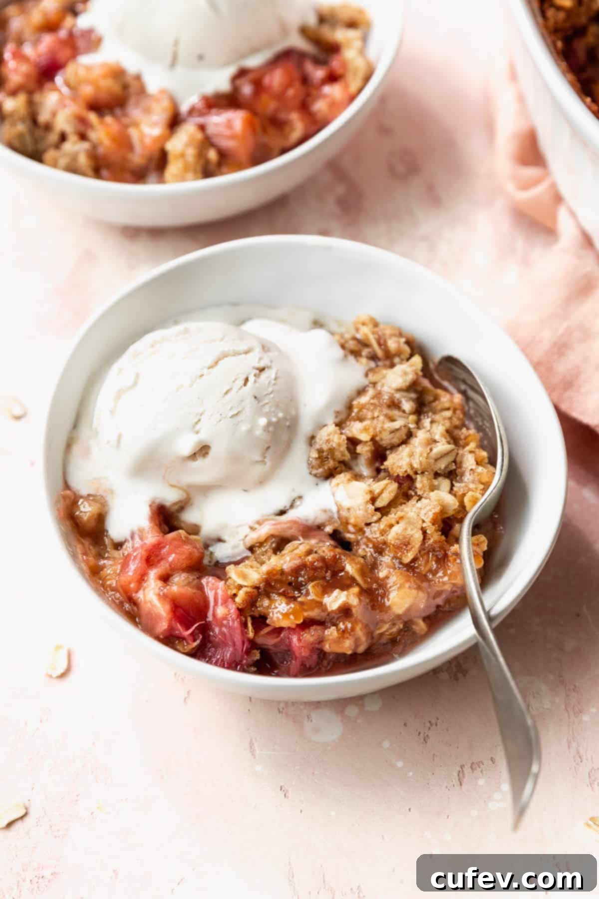 Rustic Rhubarb Crisp Gluten Free 9 45 degree angle shot of a white bowl filled with gluten-free rhubarb crisp with oats crumble topping and a scoop of vanilla ice cream on a pink table.