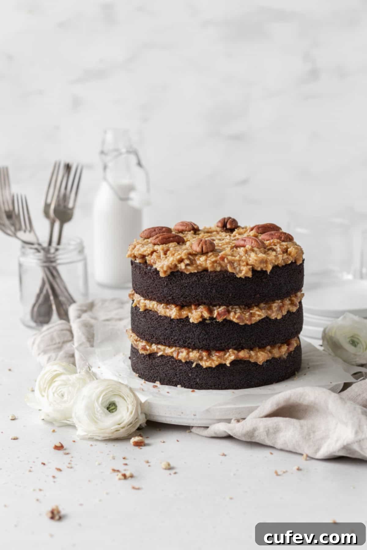 A three-layer gluten-free German chocolate cake on a round wooden board with white flowers beside it, with a milk jug and a jar with forks inside in the background.