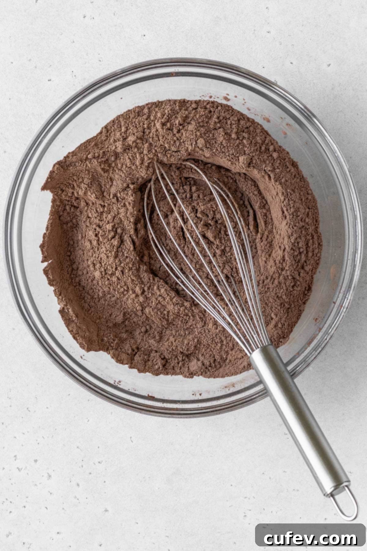 Dry ingredients for a gluten-free chocolate cake in a glass bowl with a silver whisk.