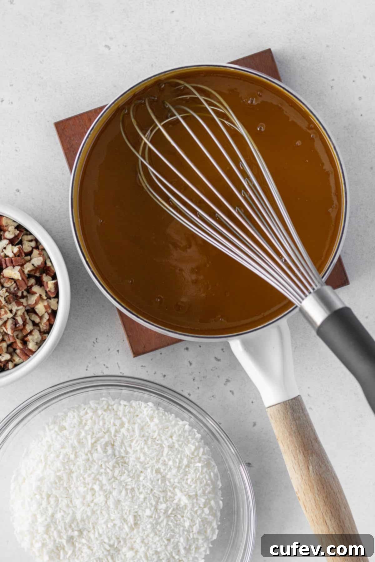 German chocolate frosting mixture being cooked in a saucepan with a bowl of shredded coconut and a bowl of chopped pecans next to it.