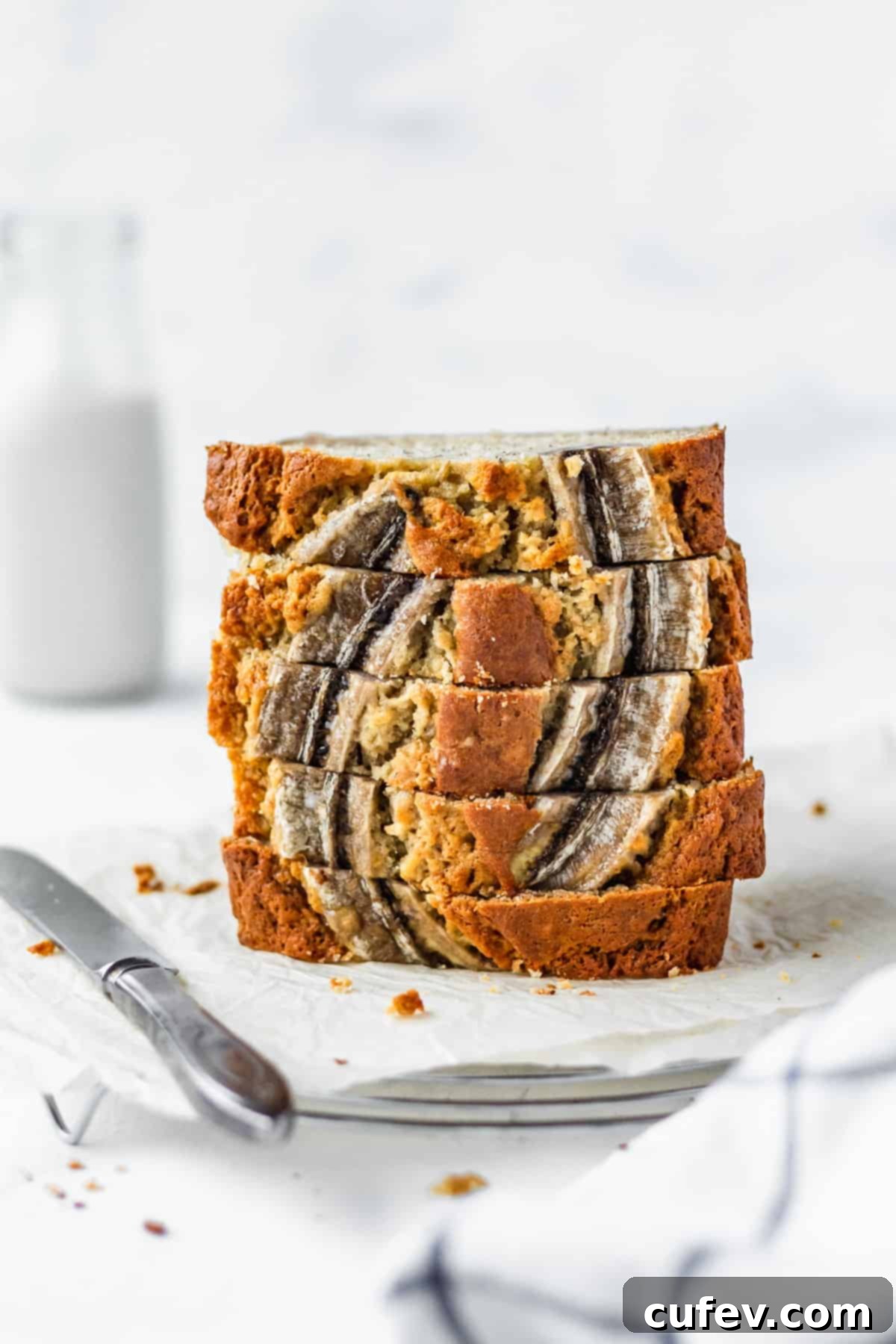 A stack of golden brown, moist banana bread slices on parchment paper, ready to be enjoyed, with a bottle of milk in the soft-focus background.