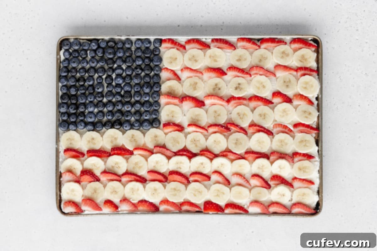 Alternating rows of neatly sliced bananas and vibrant strawberries have been added, perfectly filling out the rest of the sheet pan frame to complete the American flag design on the fruit cookie pizza.