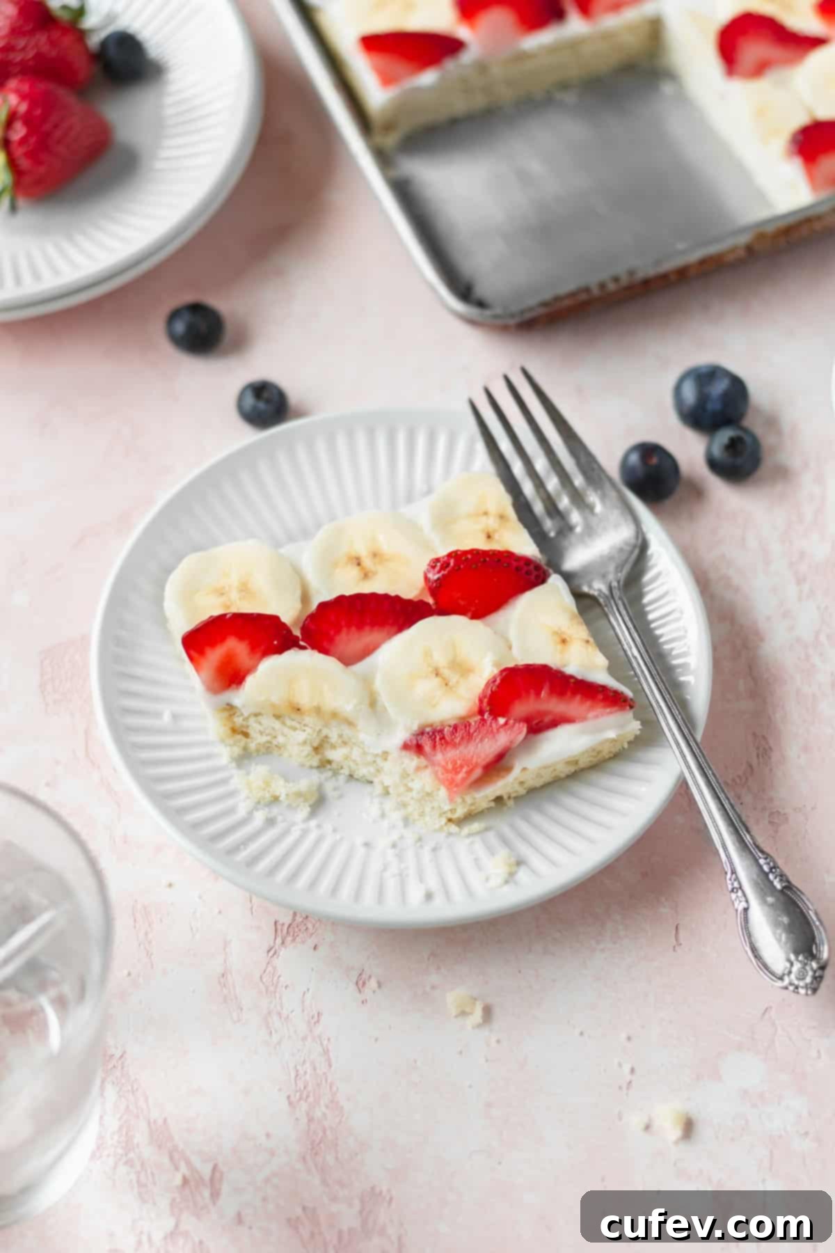 A perfectly presented slice of American flag fruit pizza, featuring vibrant blueberries, strawberries, and bananas, resting elegantly on a white dessert plate with a gleaming silver fork beside it.
