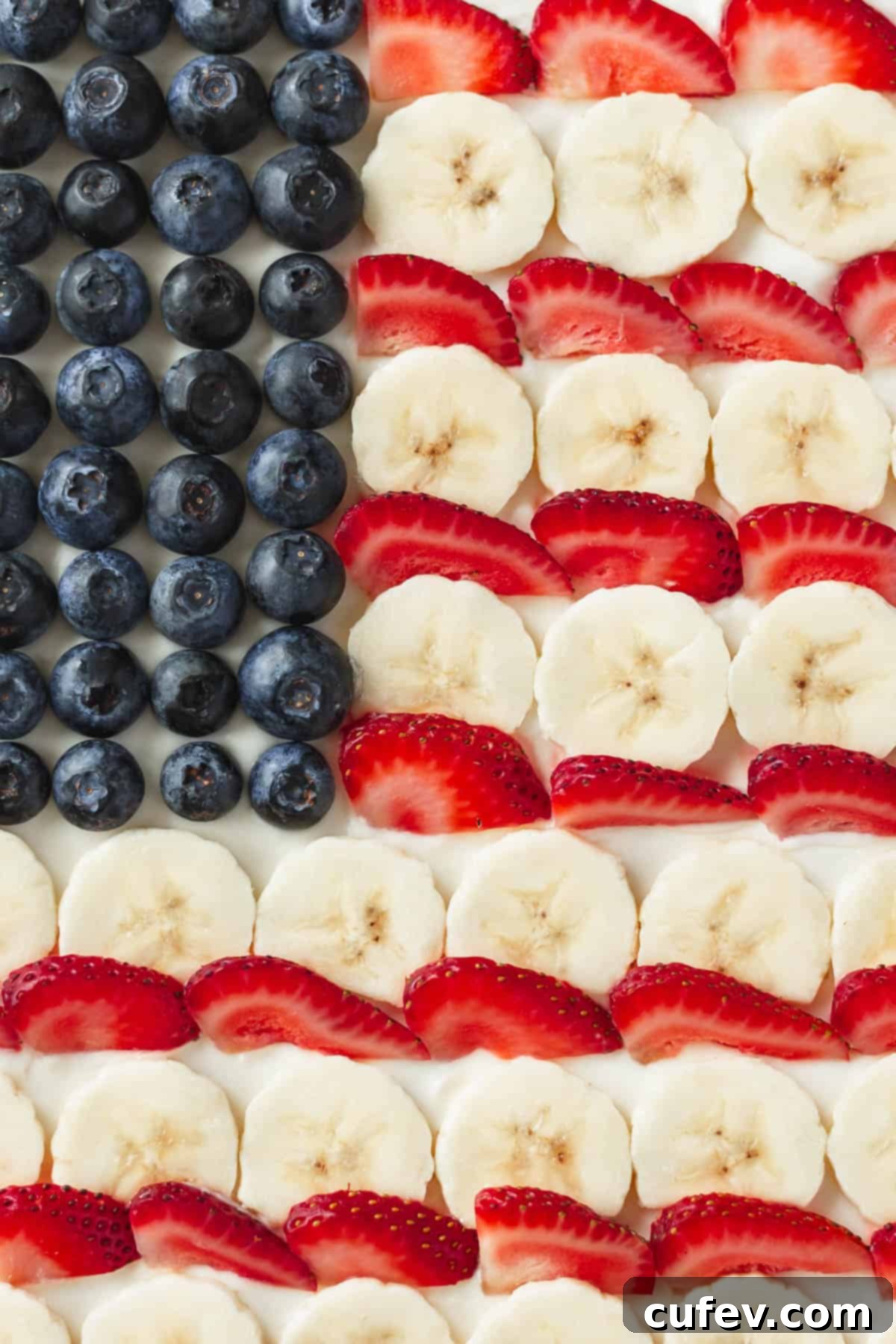 A close-up view of blueberries, perfectly sliced bananas, and vibrant sliced strawberries meticulously arranged like the American flag on a soft sugar cookie "pizza" base.