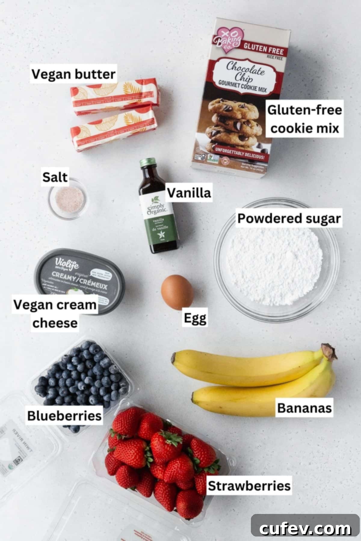 A vibrant flat lay of all the essential ingredients needed for making a delicious sugar cookie fruit flag dessert pizza, neatly arranged on a crisp white table, with an informative text overlay.
