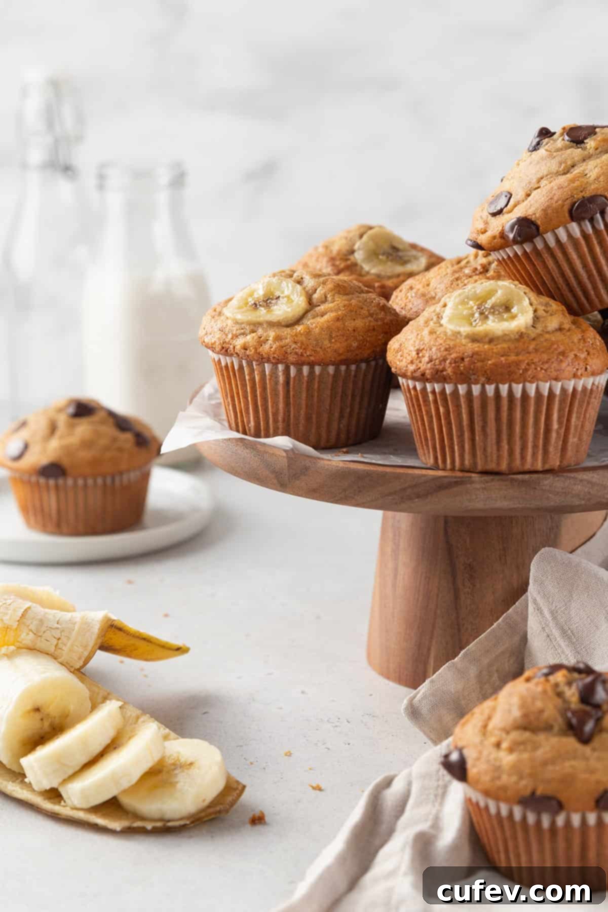 Fluffy gluten-free banana muffins cooling on a wooden cake stand, with sliced bananas and glass milk bottles in the background.