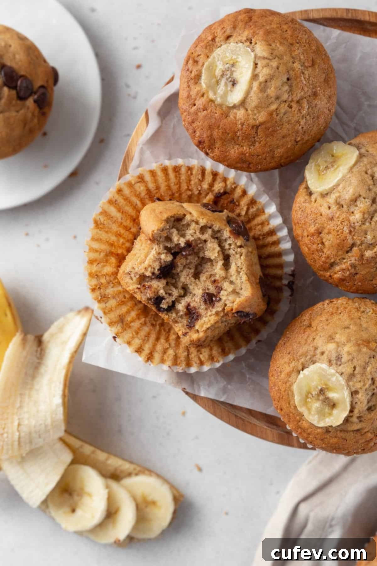 A close-up of a bitten chocolate chip banana muffin in its paper liner, surrounded by three more freshly baked muffins.