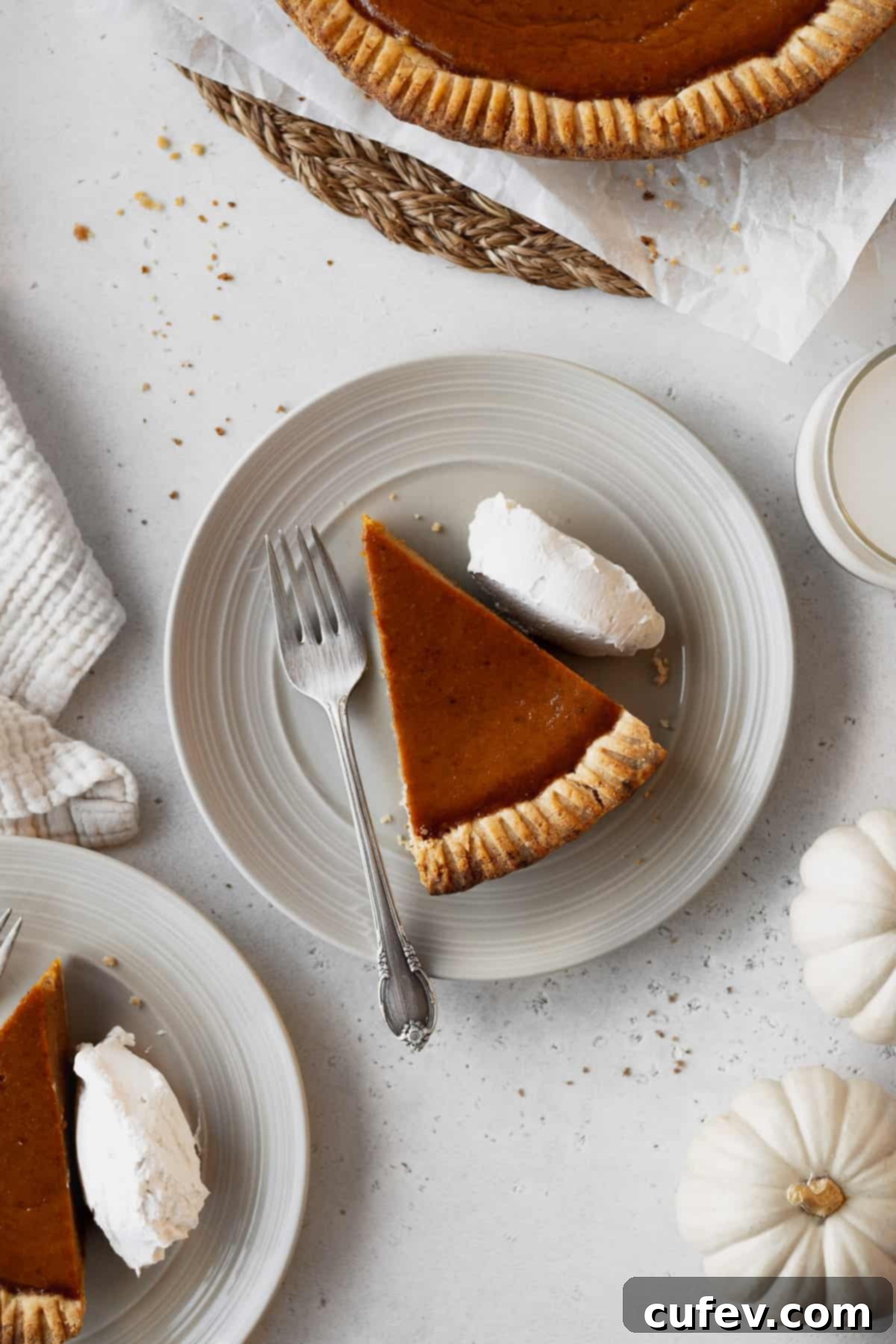 An inviting overhead shot of a slice of dairy-free pumpkin pie topped with whipped cream, accompanied by a fork on a plate, surrounded by other plates and charming white pumpkins, perfect for a festive autumn table.